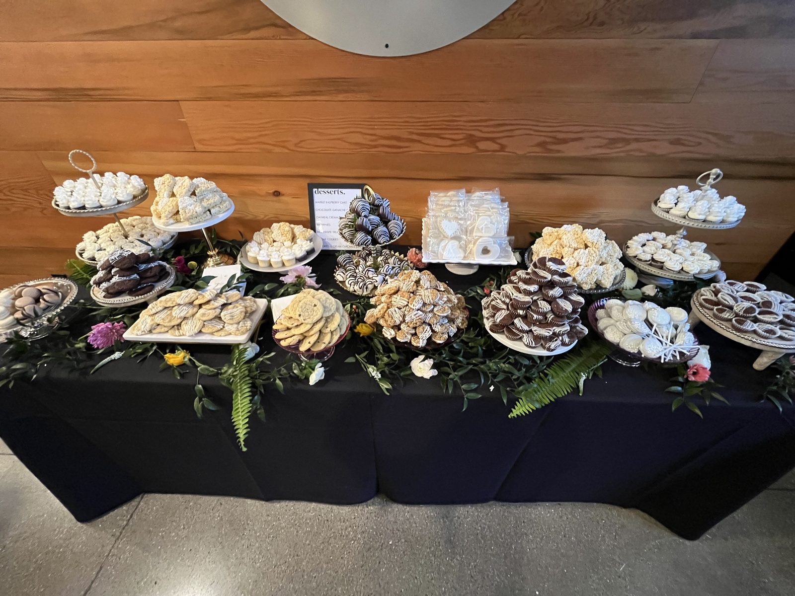 Eat Cute Bakery dessert table at a wedding — tiered stands of cupcakes, krispies, cookies, and chocolate-dipped treats with greenery and flowers